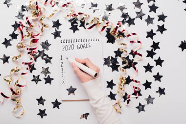 cropped view of businesswoman writing 2020 goals list in notebook near decorative stars and serpentine on white table clipart