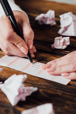 partial view of gambler marking numbers in lottery ticket near crumpled lottery cards on wooden table clipart