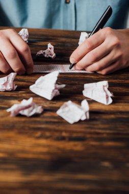 cropped view of gambler marking numbers in lottery ticket near crumpled lottery cards on wooden table clipart