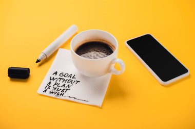 Kâğıt peçeteli kahve fincanı ve plansız bir şekilde sadece yazı, akıllı telefon ve sarı masadaki keçeli kalem.