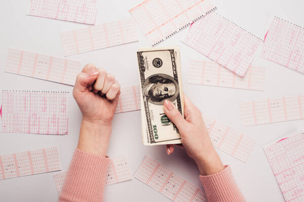 cropped view of woman showing winner gesture while holding dollar banknotes near lottery tickets scattered on white table