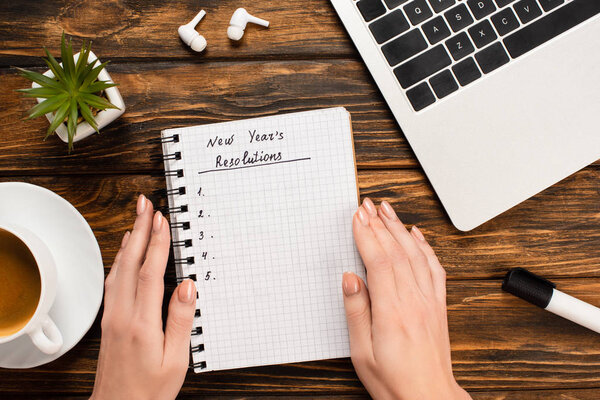 partial view of female hands near notebook with new years resolutions lettering near laptop, wireless earphones, plant and coffee cup on wooden desk