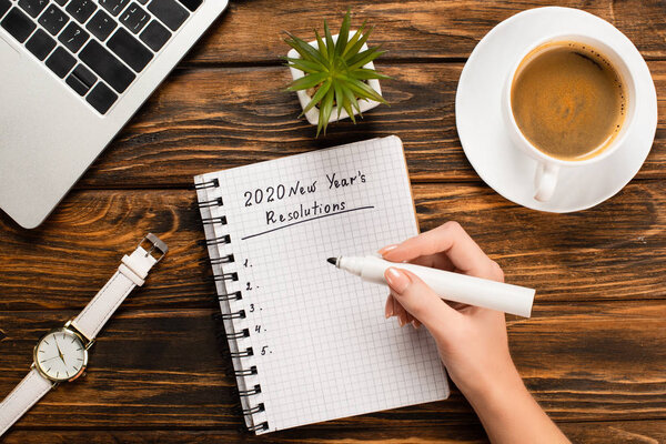 cropped view of businesswoman holding felt-tip pen near notebook with 2020 new years resolution near coffee cup, laptop, wristwatch and potted plant on wooden desk