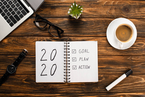 notebook with 2020, goal, plan, action lettering, laptop, coffee cup, felt-tip pen, glasses, wristwatch and plant on wooden desk