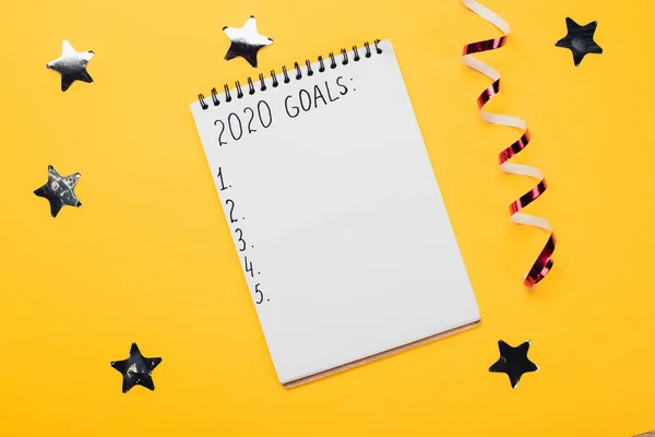 notebook with 2020 goals inscription with empty numbered points near decorative, shiny stars and serpentine on yellow surface