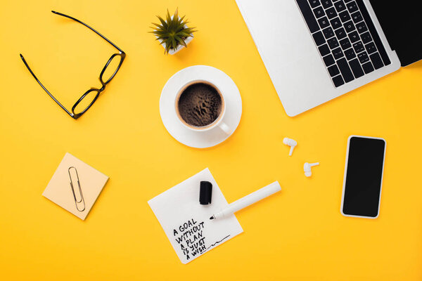 paper napkin with goal without plan just wish inscription, felt-tip pen, laptop, smartphone, coffee cup, glasses, wireless headphones on yellow desk