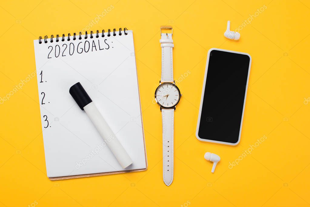 notebook with 2020 goals inscription and empty numbered points near smartphone, wristwatch and wireless earphones on yellow surface