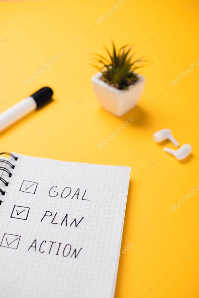 notebook with goal, plan, action words near potted plant, wireless earphones and felt-tip pen on yellow desk