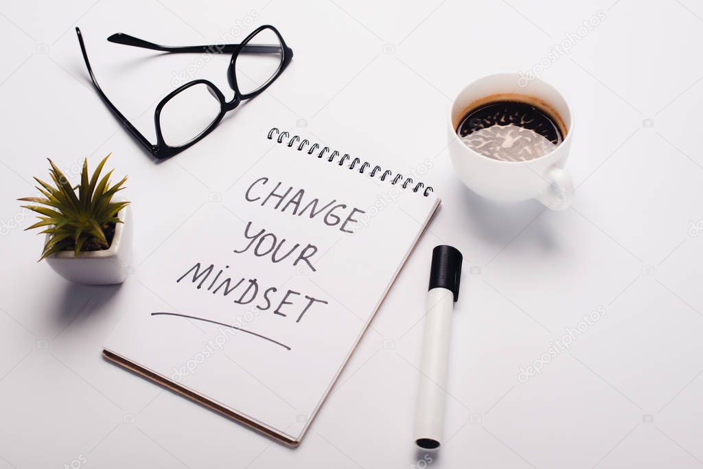 notebook with change young mindset inscription, felt-tip pen, coffee cup, glasses and potted plant on white surface