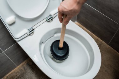 cropped view of plumber using plunger in toilet bowl in modern restroom with grey tile clipart