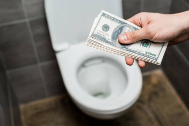 Modern tuvaletteki gri fayanslı klozetin yanında dolar banknotları tutan adam manzarası.