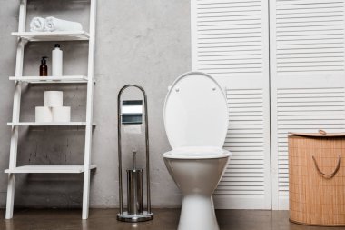 interior of modern bathroom with toilet bowl near rack with cosmetics, towels, toilet paper, laundry basket and toilet brush clipart