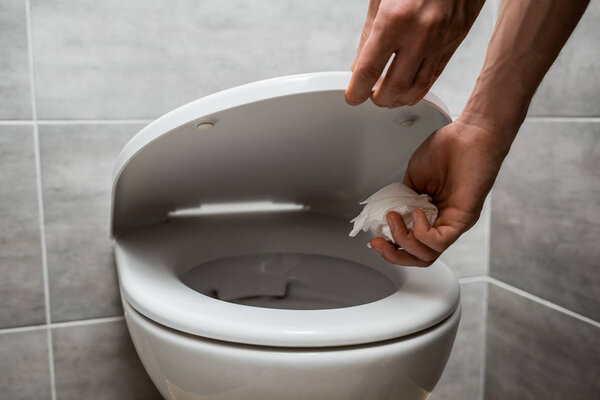 cropped view of man holding toilet lid and throwing paper in toilet bowl in modern restroom with grey tile