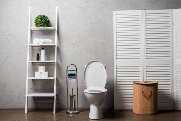 interior of modern bathroom with toilet bowl near rack with cosmetics, towels, toilet paper, laundry basket and toilet brush