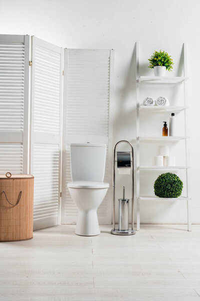 interior of white modern bathroom with toilet bowl near folding screen, laundry basket, rack and plants
