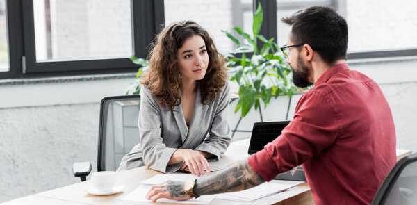 panoramic shot of account manager sitting at table and looking at colleague 