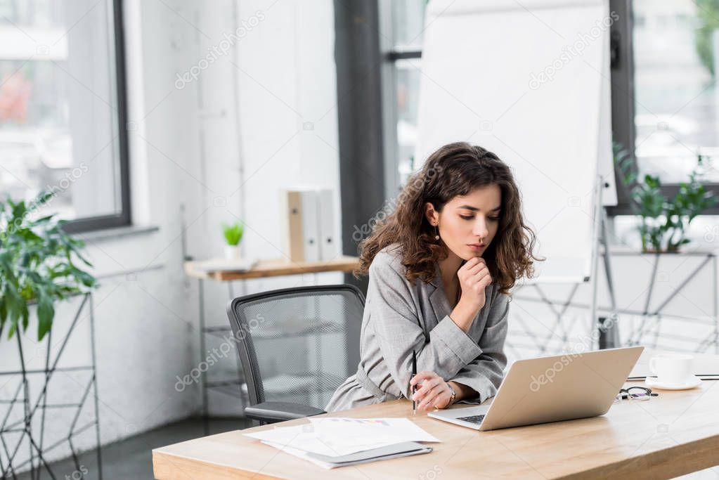 pensive account manager sitting at table and looking at laptop 