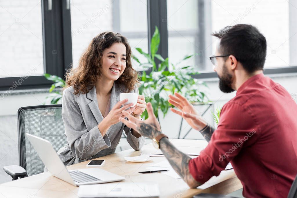 smiling account manager holding cup and talking with colleague 