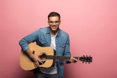 Genç adam kameraya gülümsüyor ve pembe arka planda akustik gitar çalıyor.