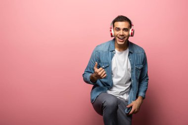 Kulaklıklı adam kameraya gülümsüyor ve pembe arka planda işaret gibi görünüyor.