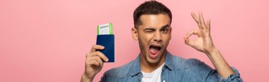 Panoramic shot of man with passport and boarding pass winking and showing okay sign isolated on pink  clipart
