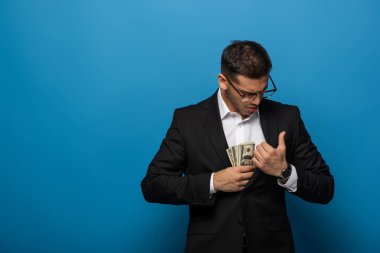 İşadamı mavi arka planda ceketin cebine dolar banknotları koyuyor