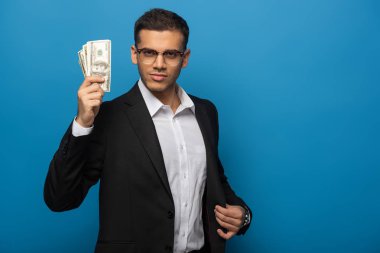 Elinde dolar banknotları olan yakışıklı bir işadamı mavi arka plandaki kameraya bakıyor.