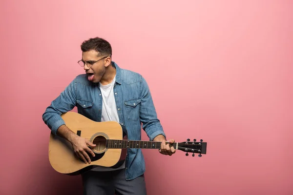 若いです男アコースティックギターを演奏中に舌を突き出すピンクの背景 — ストック写真