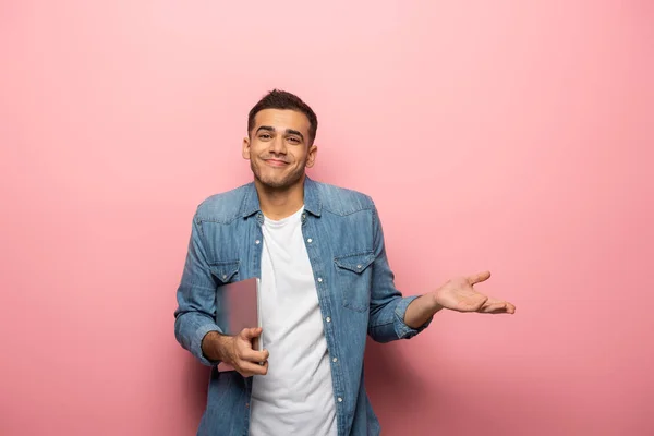 Mladý Usměvavý Muž Notebookem Ukazuje Pokrčení Rameny Gesto Růžovém Pozadí — Stock fotografie