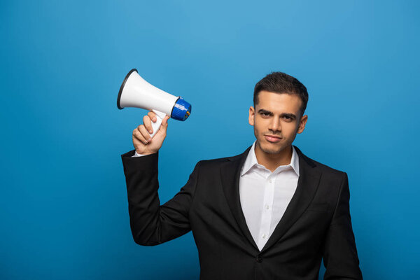 Красивый бизнесмен смотрит в камеру, держа громкоговоритель на синем фоне
