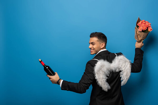 Back view of smiling businessman with white wings holding wine bottle and bouquet on blue background