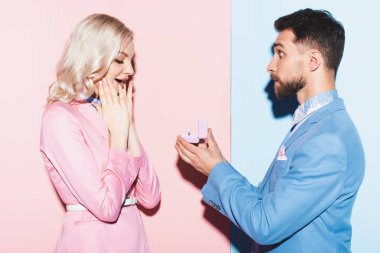 Yakışıklı bir adam pembe ve mavi arka planda şok olmuş bir kadına evlenme teklif ediyor. 
