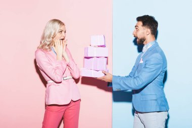 Yakışıklı adam, pembe ve mavi arka plandaki şok olmuş kadına hediyeler veriyor. 
