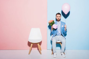 Yakışıklı ve üzgün adam elinde balon, hediye ve buket mavi ve pembe arka planda 