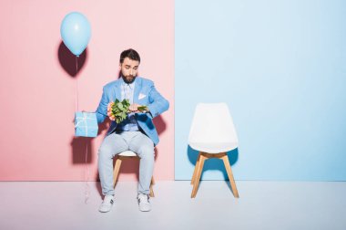 Yakışıklı ve gergin bir adam kol saatine bakıyor ve elinde balon, hediye ve mavi ve pembe arka planda 