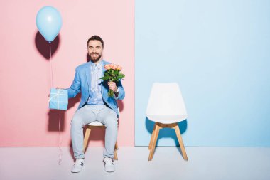 Yakışıklı ve gülümseyen adam kol saatine bakıyor ve elinde balon, hediye ve mavi ve pembe arka planda 