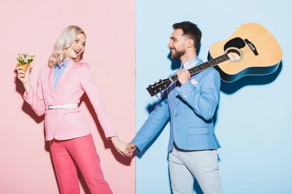 smiling woman with bouquet and handsome man with acoustic guitar holding hands on pink and blue background