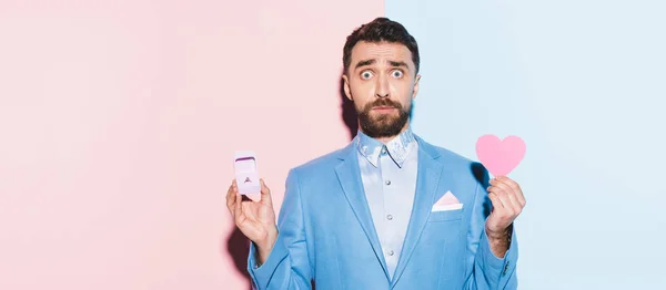 stock image panoramic shot of scared man holding heart-shaped card and engagement ring on blue and pink background 