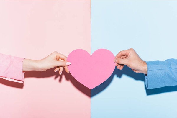 cropped view of man and woman holding heart-shaped card on pink and blue background 