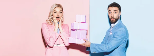 ピンクと青の背景でショックを受けた女性に贈り物をするハンサムな男のパノラマ写真 — ストック写真