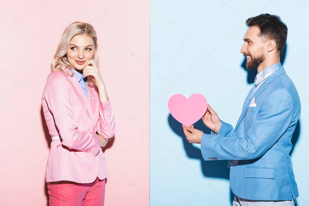 handsome man giving heart-shaped card to dreamy woman on pink and blue background 