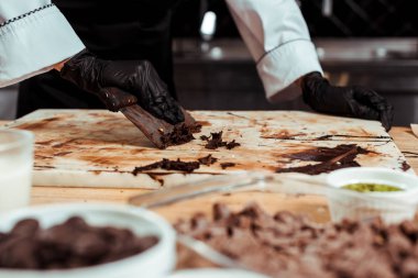 cropped view of chocolatier in black latex gloves scraping dark chocolate from surface  clipart