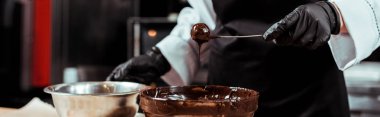 panoramic shot of chocolatier in black apron holding stick with tasty candy near chocolate in bowl  clipart