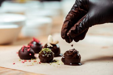 cropped view of chocolatier in black latex glove adding chocolate shavings on fresh made candies  clipart