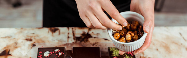 panoramic shot of chocolatier holding bowl with caramelized hazelnuts near chocolate bars 