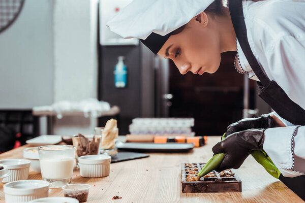 side view of chocolatier in latex gloves holding pastry bag with caramelized nuts near chocolate molds 