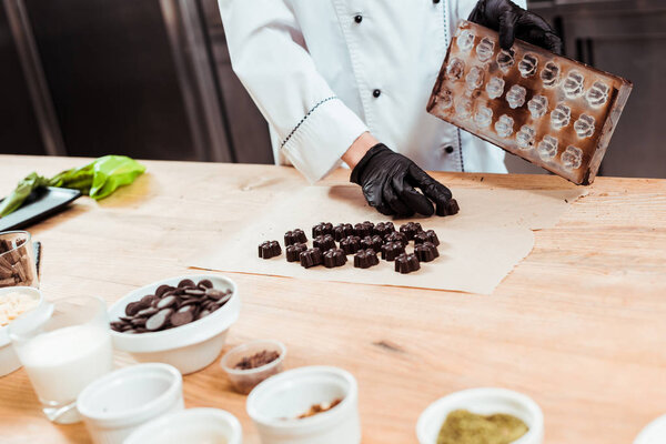 cropped view of chocolatier holding ice tray near prepared chocolate candies on baking paper 