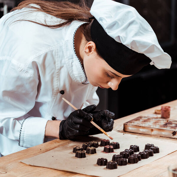 attractive chocolatier holding brush with gold powder near prepared chocolate candies on baking paper 