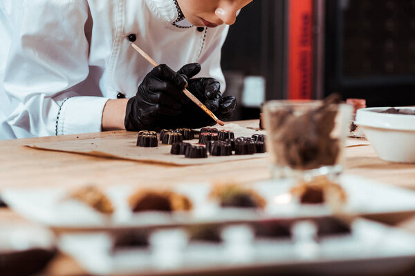 selective focus of chocolatier holding brush with gold powder near prepared chocolate candies on baking paper 
