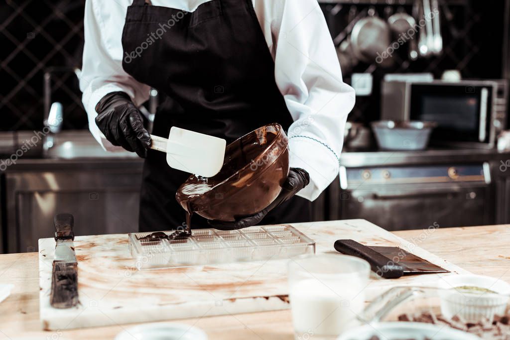 cropped view of chocolatier in apron pouring melted chocolate into ice tray 